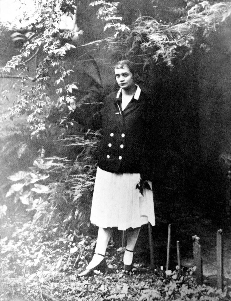 Fotografía en blanco y negro de una joven de pie entre plantas, con chaqueta oscura y falda clara.